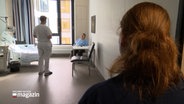Eine Person befragt einen Patienten im Krankenhaus. In der Tür zum Zimmer steht eine weitere Person die die die Szenerie beobachtet. © NDR Foto: NDR Screenshots