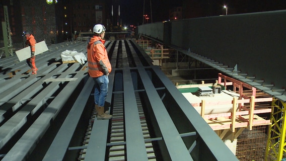 Ein Bauarbeiter steht beim Brückenbau in Lübeck vor einem schwebenden Stahlträger © NDR 