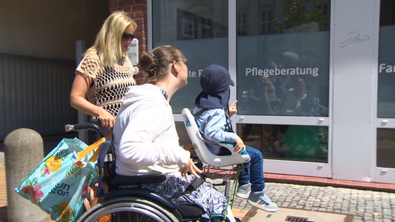 Eine Frau sitzt im Rollstuhl mit einem befestigten Multifunktionsadapter, in dem ein Kleinkind sitzt © NDR Foto: NDR