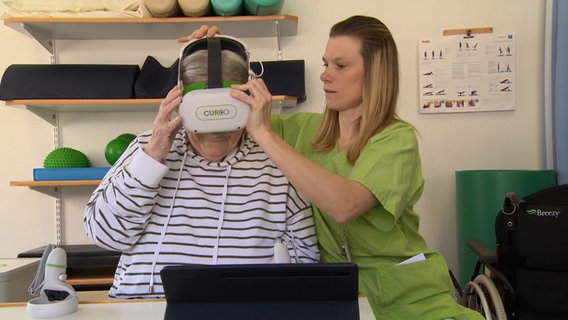 Eine Patientin bekommt von einer Physiotherapeutin eine VR-Brille aufgesetzt © NDR Foto: NDR Screenshots