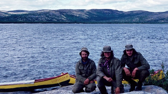 Auf einem alten Foto sitzt Arved Fuchs (M.) mit zwei Freunden an einem Ufer. Im Hintergrund liegen Kanus im Wasser. © Screenshot Foto: NDR