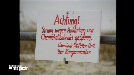 Eine historische Aufnahme zeigt ein weißes Warnschild mit dem Text in rot an einem Strandabschnitt in St. Peter-Ording. Der Text auf dem Schild lautet Achtung! Strand wegen Anlandung von Chemikalienbeutel gesperrt. Gemeinde St. Peter-Ord. Der Bürgermeister. © NDR 