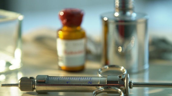 Eine Spritze und Medikamente liegen auf einem Tisch. © NDR 