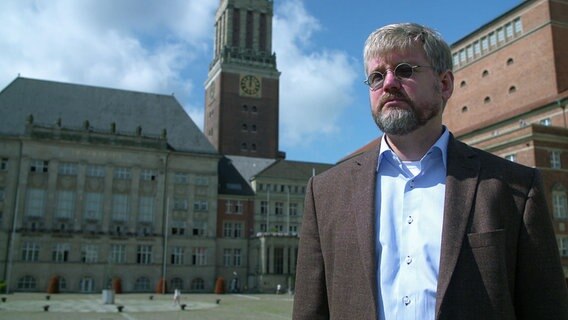 Ein Mann von der Kieler Stadtplanung steht vor dem Rathausplatz am alten Kieler Rathaus. Er trägt eine kleine runde Sonennbrille © NDR / SH Magazin 