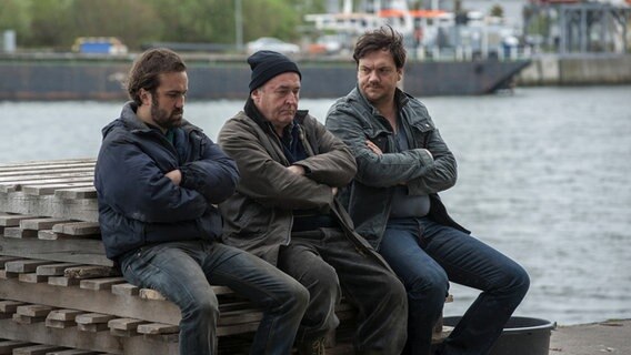 Kommissar Bukow (Charly Hübner) sitzt mit zwei Männern auf gestapelten Paletten im Hafen. © NDR/Christine Schroeder 