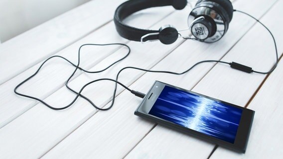 Handy mit angeschlossenen Kopfhörern auf einem weißen Tisch. Auf dem Display ist eine blaue Audio-Wellenform zu sehen. © IMAGO Foto: agefotostock