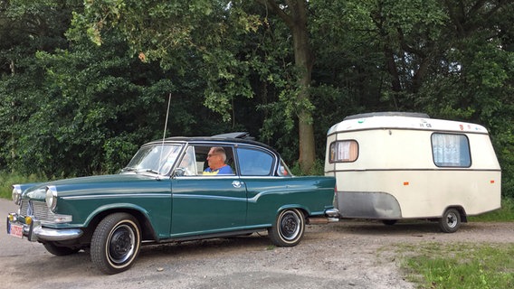 Ein petrolfarbenes Auto aus den 1960er-Jahren zieht einen kleinen weißen Wohnwagen aus den 1960er-Jahren. © NDR/Lona Media GbR 