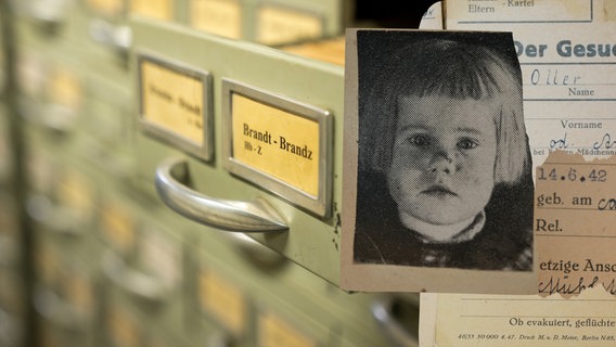Eine Karteikarte eines vermissten Kindes vor einem Aktenschrank mit einer geöffneten Schublade. © NDR 