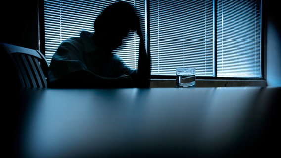 Eine Person sitzt mit auf den Händen gestütztem Kopf in einem dunklen Raum am Tisch. © DNY59 / iStockphoto Foto: DNY59