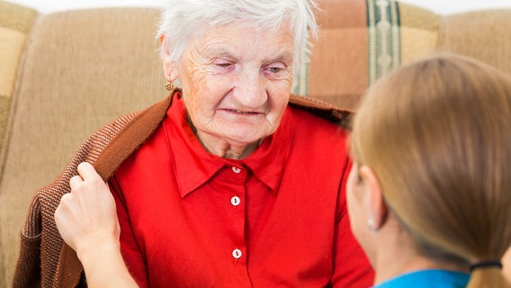 Pflegerin legt eine Jacke um eine Seniorin, die auf einem Sofa sitzt. © Fotolia.com Foto: Ocskay Bence