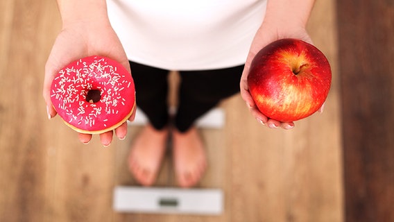 Frau steht auf einer Waage und hält einen Doughnut und einen Apfel in den Händen © Fotolia.com Foto: Maksymiv Iurii