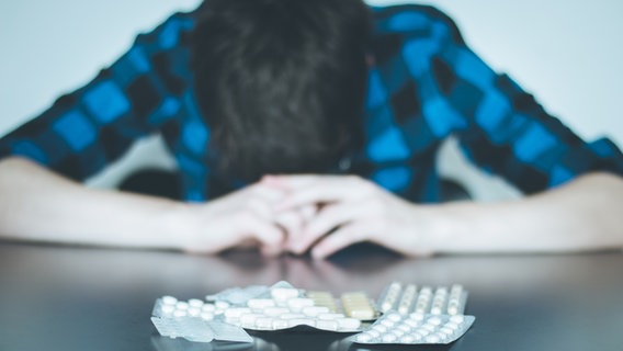 Varios medicamentos están sobre la mesa frente a un hombre desesperado.  © fotolia Foto: Patrick Daxenbichler
