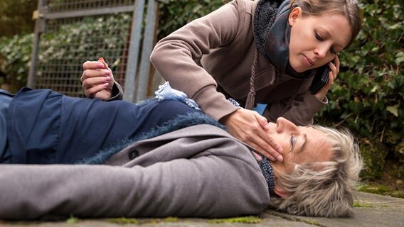 Eine ältere Frau liegt mit geschlossenen Augen am Boden, eine junge Frau beugt sich zu ihr und ordert telefonisch Hilfe © fotolia.com Foto: M.Dörr & M.Frommherz