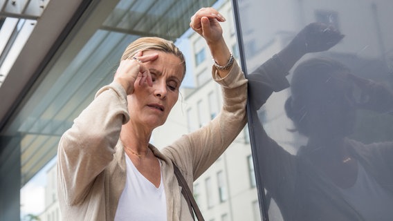 Eine Frau greift sich im Stehen an die Stirn, stützt sich an der Wand ab und schließt die Augen. © picture alliance Foto: Christin Klose