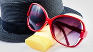 Ein Sonnenhut, eine Sonnenbrille und Sonnenschutzcreme liegen auf einem Tisch. © Colourbox 