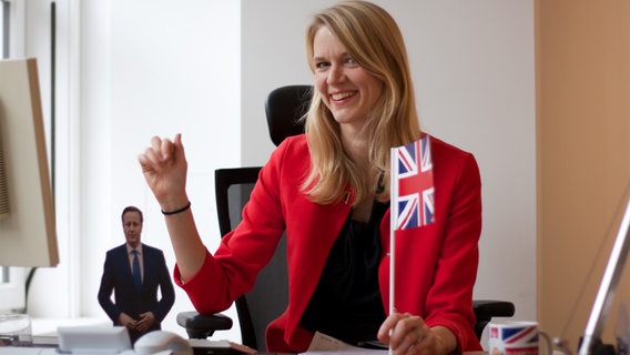 ARD Korrespondentin Julie Kurz an einem Schreibtisch mit Union Jack und einer Aufstellfigur von David Cameron.  