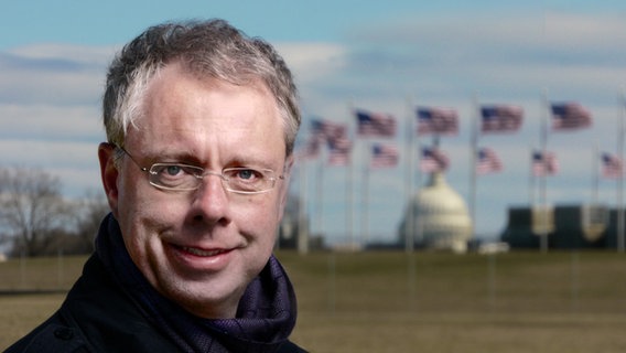 ARD-Korrespondent Stefan Niemann steht vor USA-Flaggen.  