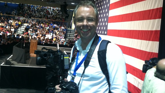 ARD-Korrespondent Stefan Niemann mit umgehängter Kamera vor einer großen USA-Fahne.  