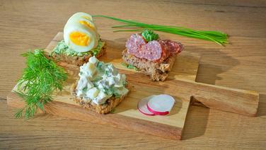 Three types of sandwiches on wholemeal bread with chicken salad, sausage and herb butter with eggs, arranged on a wooden board.  © NDR/doclights Photo: Marc Vorwerk