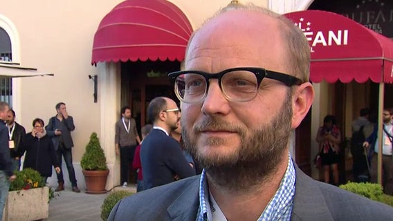 "Correctiv"-Herausgeber David Schraven (48) wird vor einem Hotel interviewt. Er trägt eine Brille und einen Vollbart, hat ein Sakko und ein kariertes Hemd an.  