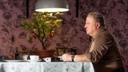 Marko Wendrichs (gespielt von Axel Prahl) sitzt an einem Esstisch. Seine Hände hat er auf der Tischplatte abgelegt und schaut starr geradeaus. © NDR/Gordon Timpen 