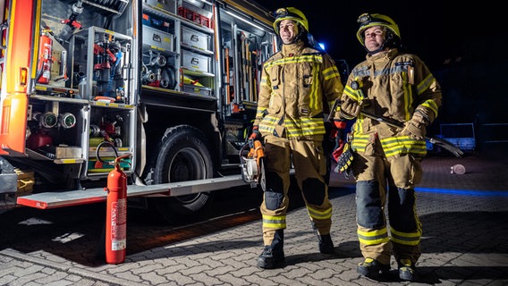 Zwei Feuerwehrmänner laufen in Montur neben einem Einsatzfahrzeug entlang. © NDR/Kamera Zwei 