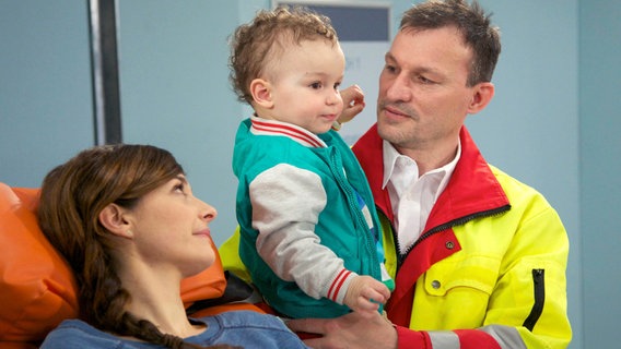 Sabine Schrader (Anne Werner, l.) wird nach einem Sturz von einer Leiter in die Sachsenklinik eingeliefert. Ihren kleinen Sohn Tim (Finn Graul, Mitte) musste der Notarzt (Komparse, r.) mitnehmen, da Sabine alleinerziehend ist. © NDR/MDR/Saxonia/Wernicke 