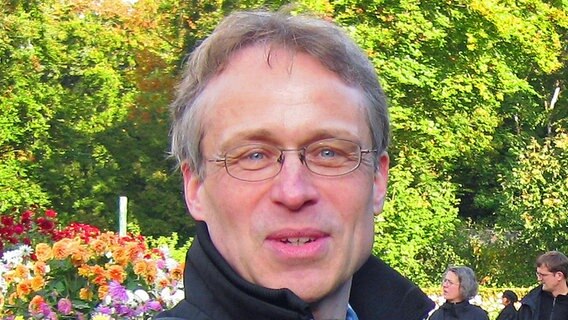 Rudolf Beckmann: ein graumelierter Mann um die 50 in einem Park. Er hat ein schmales Gesicht, blaue Augen und trägt eine kleine Brille.  
