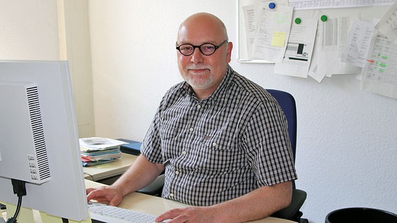 Peter Lindner: ein Mann um die 50, der in einem Büro an einem PC sitzt und in die Kamera lächelt. Er hat einen grauen Haarkranz, einen grauen Bart und trägt ein braun-weiß kariertes Hemd und eine schwarze Hornbrille.  Foto: Martin Ovelgönne, NDR