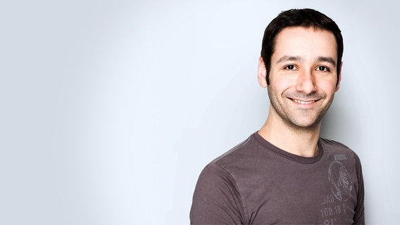 Tarek Youzbachi: ein Mann um die 40 mit kurzen schwarzen Haaren und dunklen Augen. Er hat einen Dreitagebart und trägt ein dunkelgraues T-Shirt. © NDR 