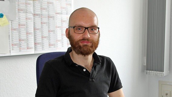 Dennis Tietz: ein Mann Anfang 30 mit kurzgeschorenen Haaren, rötlichem Vollbart, Hornbrille und schwarzem Polohemd in einem Büro.  Foto: Martin Ovelgönne, NDR