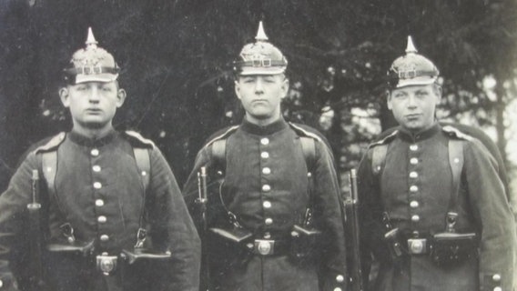 Drei jugendliche Soldaten in Uniformen mit Pickelhaube auf einer schwarzweißen Fotografie aus dem Ersten Weltkrieg.  