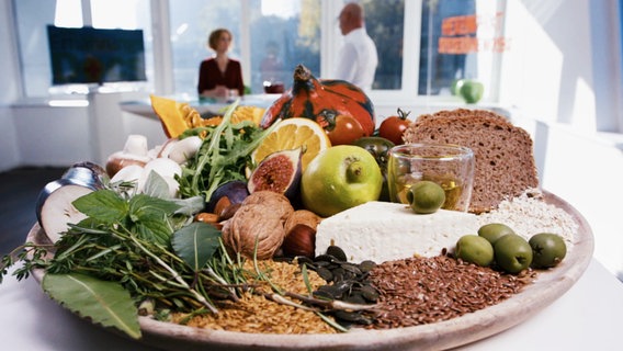 Eine PLatte mit viel Gemüse, Nüssen, Vollkornbrot und Käse steht im Vordergrund, im Hintergrund Doc Andresen und ihr Patient. © Screenshot 