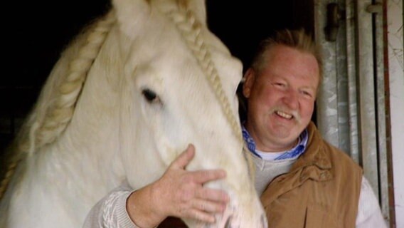 Tamme Hanken neben einem großen weißen Pferd mit geflochtener Mähne.  