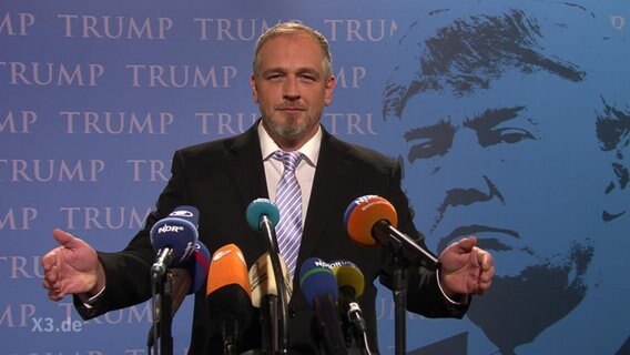 Ein Mann spricht in mehrere Mikrophone, im Hintergrund mehrfach der Schriftzug: "Trump".  