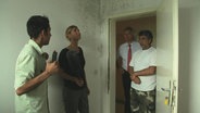 Eine Reporterin mit jungen Männern in einem von Schimmel befallenen Raum.  
