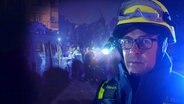 Timo Eilhardt mit Helm und Schutzkleidung, im Hintergrund sind Mitglieder seines THW-Teams beim Einsatz im Krisengebiet zu sehen. © NDR 