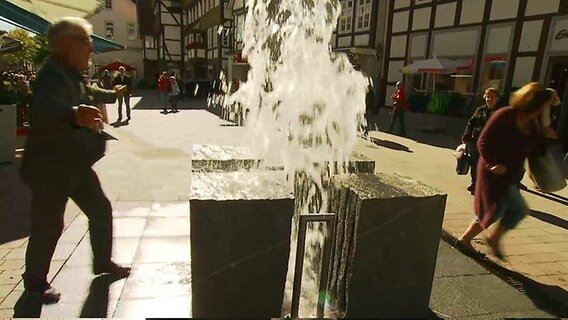 Zwei Menschen flüchten vor einer Wasserfontäne in einer Fußgängerzone.  