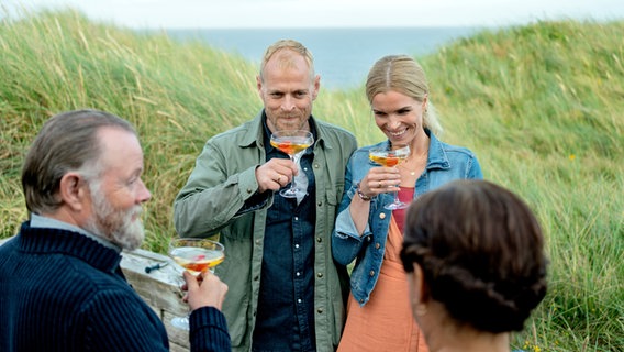 Szenenbild aus der dänischen Serie "Whits Sands": Zwei Paare stehen sich gegenüber und prosten sich zu. Im Hintergrund ist die Nordsee zu sehen. © Martin Dam Kristensen 