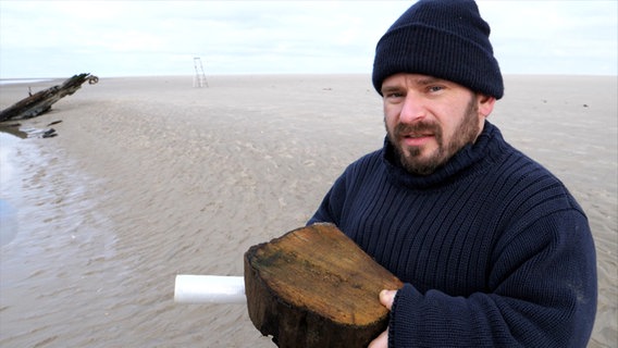 Der Archäologe Daniel Zwick hat ein Holzstück in der Hand, hinter ihm ist das Wrack eines Schiffes zu sehen. Der Archäologe Daniel Zwick hat ein Holzstück in der Hand, hinter ihm ist das Wrack eines Schiffes zu sehen. © Screenshot