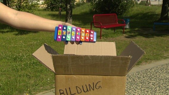 Eine Hand legt ein Kinderxylophon in einen Karton mit der Aufschrift "Bildung". Eine Hand legt ein Kinderxylophon in einen Karton mit der Aufschrift "Bildung".