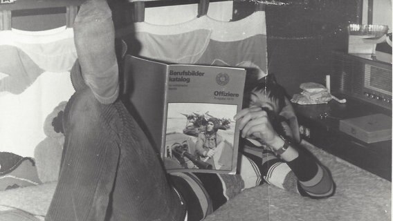 Schwarz-weiß-Foto: Ein Junge liegt auf einem Bett und liest den Berufsbilder-Katalog der NVA.  