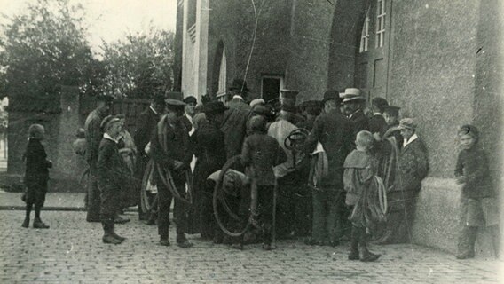 Schwarz-Weiß-Foto: Menschen stehen vor einem Haus und haben Reifenschläuche in der Hand.  