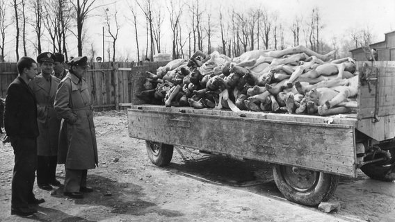 Befreiter Häftling des KZ Buchenwald mit amerikanischen Rundfunkjournalisten vor einem mit Leichen beladenen Lkw-Anhänger im Innenhof des Krematoriums 1945. © National Archives, Washington / Public Domain 