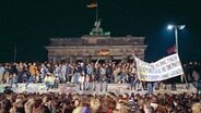 Einen Tag nach der Grenzöffnung in Berlin feiern Tausende Menschen auf, vor und hinter der Berliner Mauer am Brandenburger Tor. Auf einem Transparent im Vordergrund steht geschrieben "Für ein Berlin ohne Mauer in einem  Deutschland ohne Panzer in einem Europa ohne Grenzen". © Peter Zimmermann Foto: Peter Zimmermann