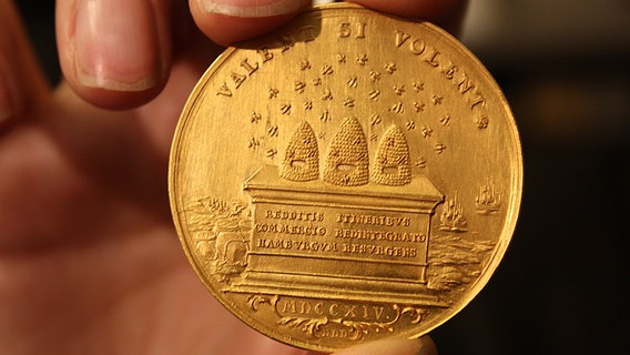 Goldmedaille von 1714 auf die Pest in Hamburg - drei Bienenkörbe und im Hintergrund Schiffe, darüber in Latein: "Die Stadt Hamburg hat ihren alten Schmuck wieder erhalten, nachdem die Einschränkungen der öffentlichen Wege aufgehoben und der Handelsverkehr wieder hergestellt worden ist." © NDR Foto: Oliver Diedrich