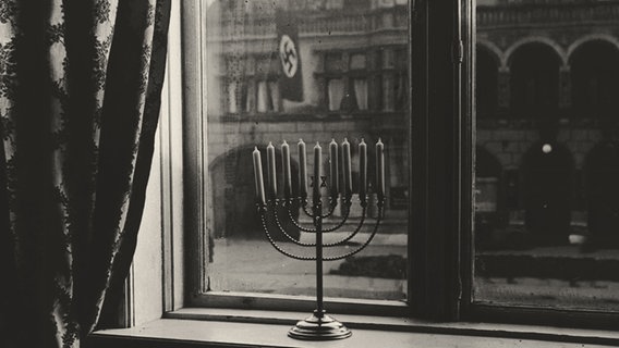 Das Foto von Rosi Rahel Posner zeigt einen Chanukka-Leuchter im Fenster. Auf der gegenüberliegenden Straßenseite liegt das Kieler Hauptquartier der NSDAP. © Nava Gilo 