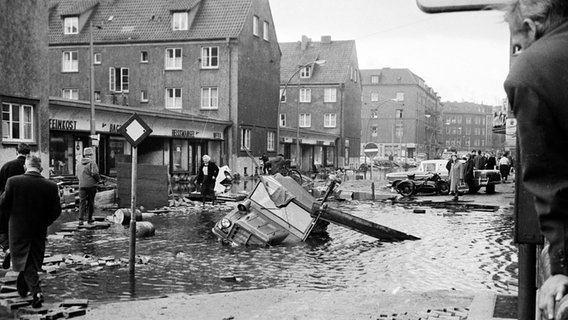 Menschen gehen an einem versunkenen DKW Munga-Geländewagen vorbei in der Einmündung und Dierksstraße in Hamburg-Wilhelmsburg nach der Sturmflut 1962. © NDR Foto: Günther Hachmeister