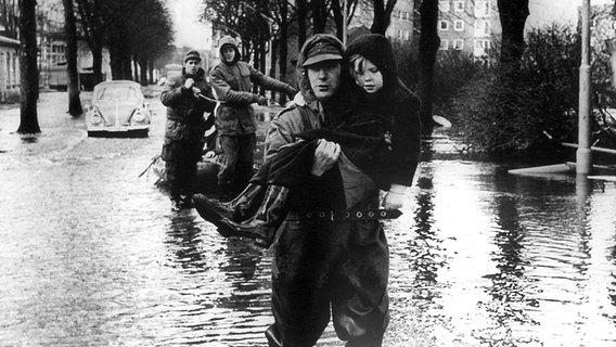 Ein Helfer trägt ein Kind über die überschwemmte Straße nach der Sturmflut 1962 © dpa / picture alliance 