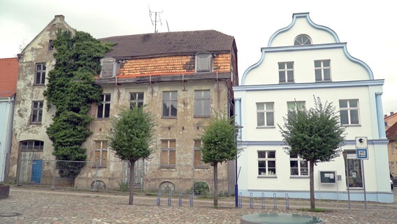 Neben einem sanierten Haus an einer Kopfsteinpflasterstraße mit Bäumchen steht eine Bauruine. © NDR 